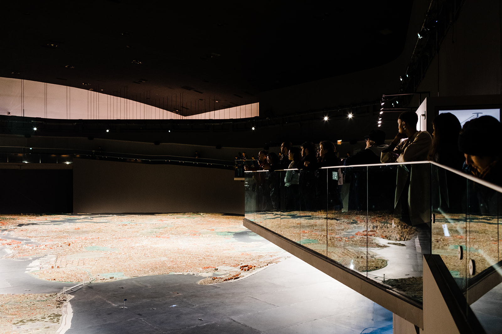 In a fairly dark room, people are standing on an overhang above the Panorama of the City of New York (a massive model that is well lit), where the assembly began.