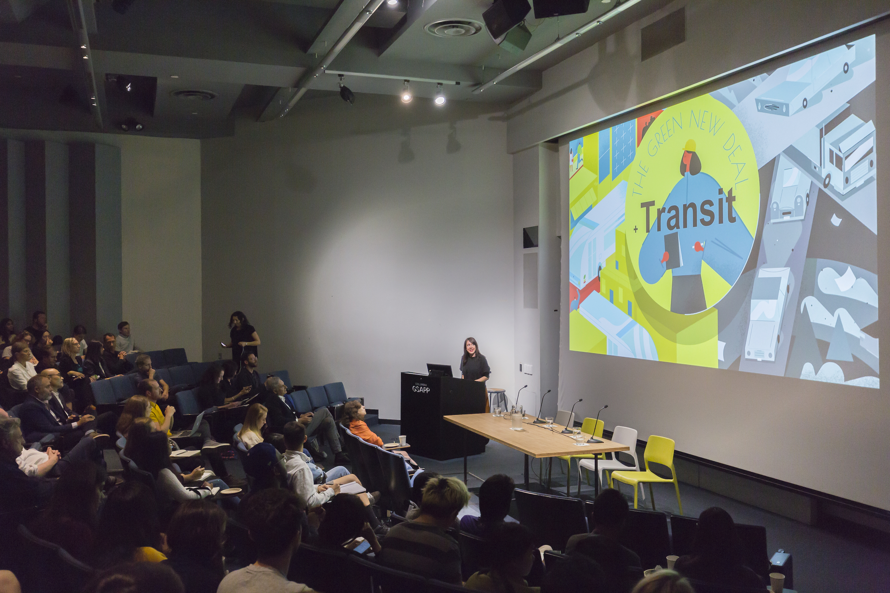 Hayley Richardson presenting on "Public Transportation" behind a podium in Wood Auditorium. Slide on projection screen reads: "The Green New Deal, Transit." 