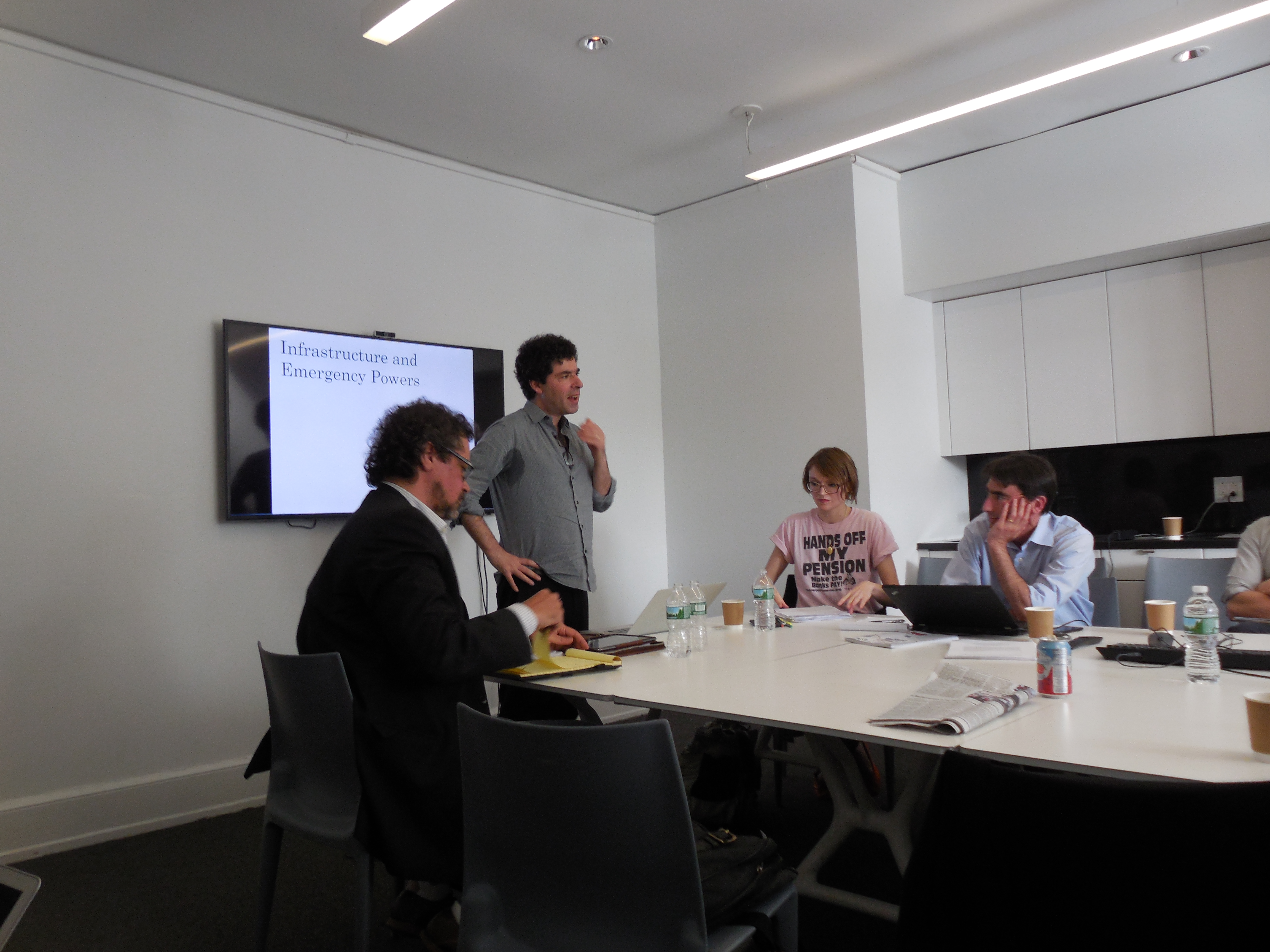 A man presenting while standing next to a screen to a group seated around a table