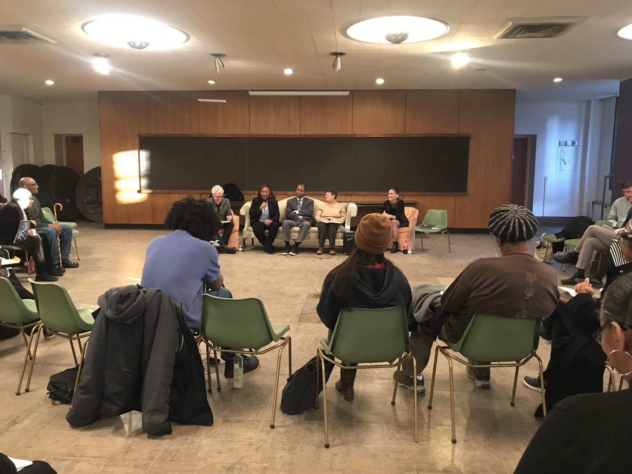 Panelists seated on a couch at the front of the room with listeners seated in a semi-circle surrounding them