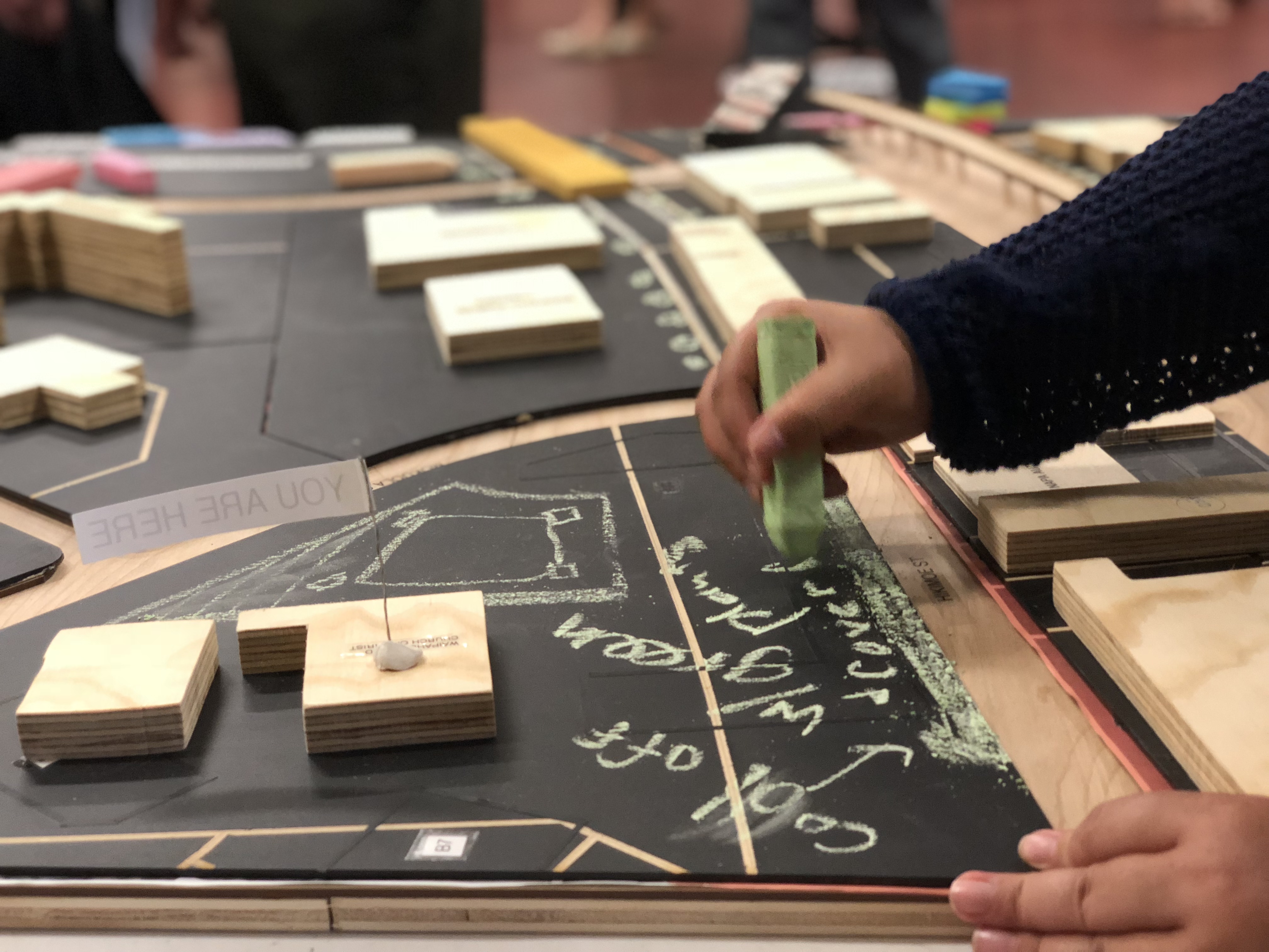A close-up image of a large-scale game made of wooden pieces, a chalkboard, and an arm extends from the right side of the image with a piece of green chalk in hand