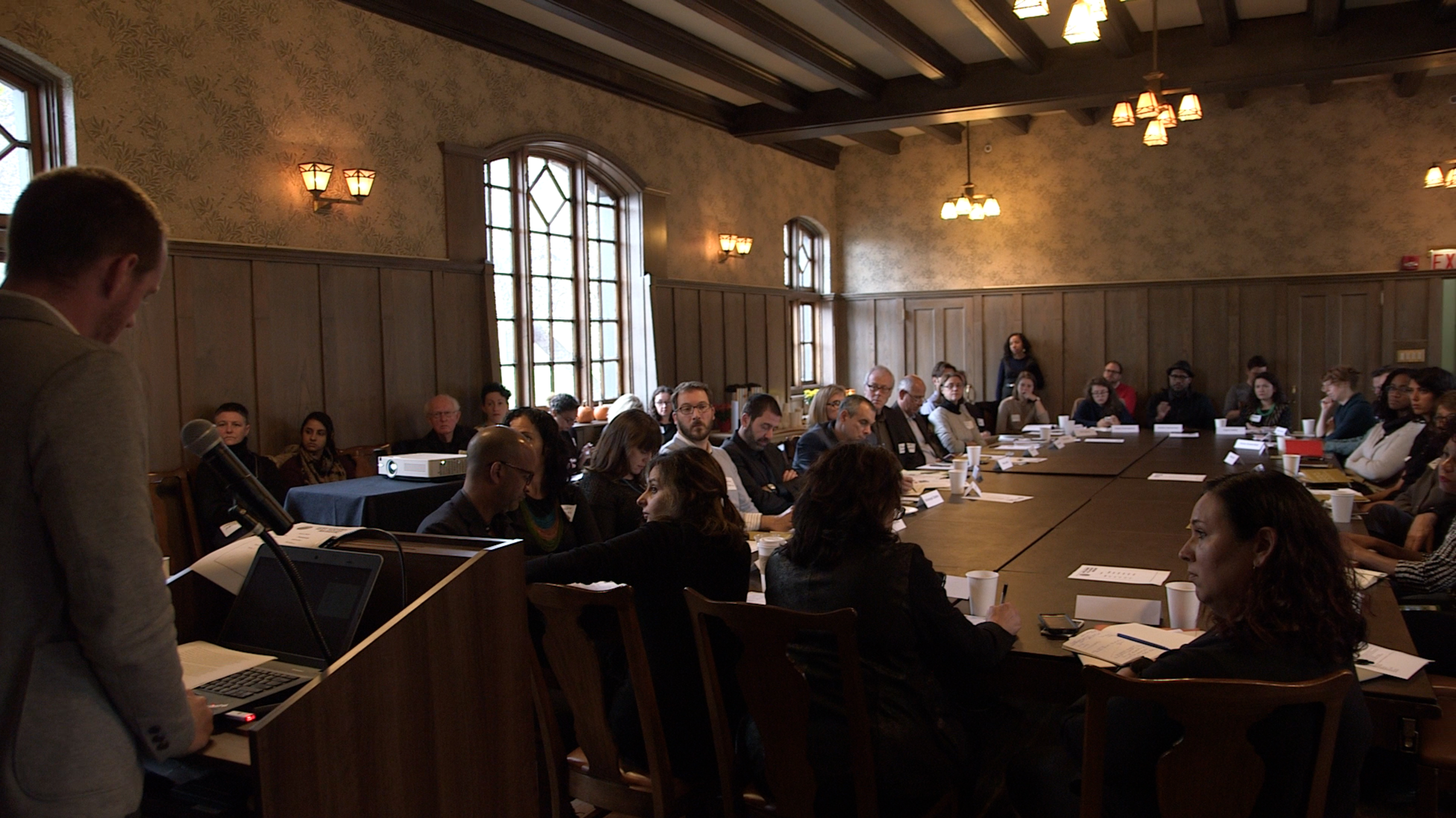A man speaking at podium in front of long table with people seated around